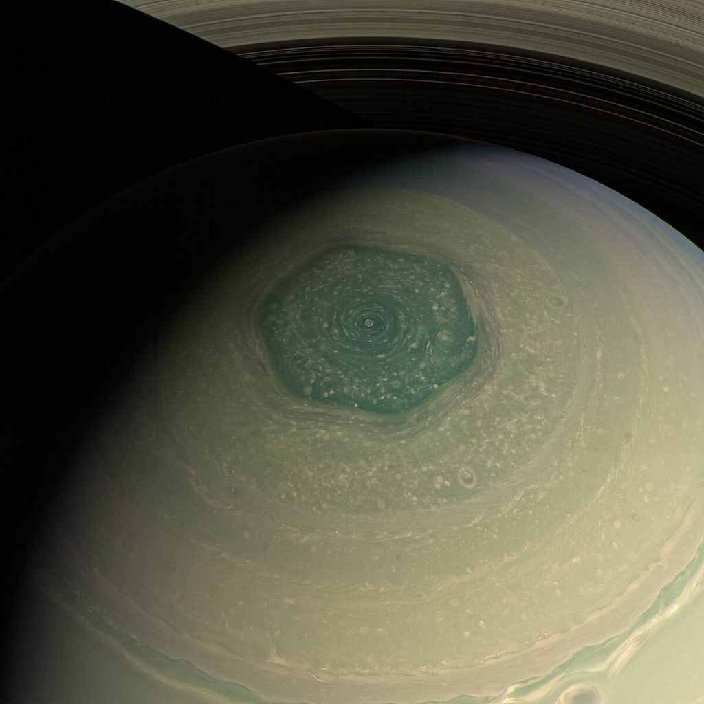 "Close-up view of Saturn's north polar hexagon, a massive six-sided jet stream surrounding a central vortex at the planet's north pole, with the rings visible at the top of the image.